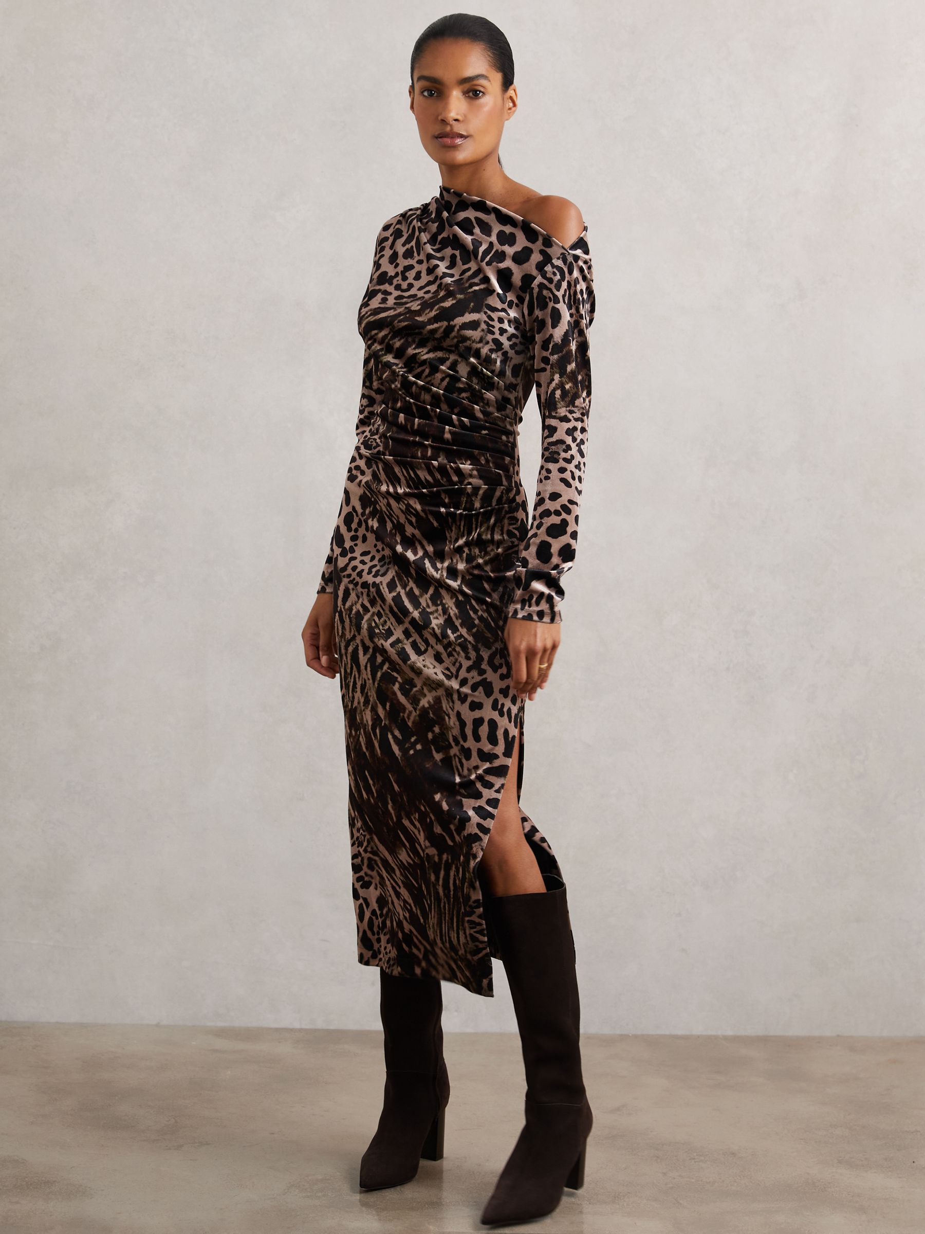 A woman wearing a velvet leopard-print midi dress with an asymmetric off-shoulder neckline, ruched waist, and high side slit, paired with black high-heeled boots.
