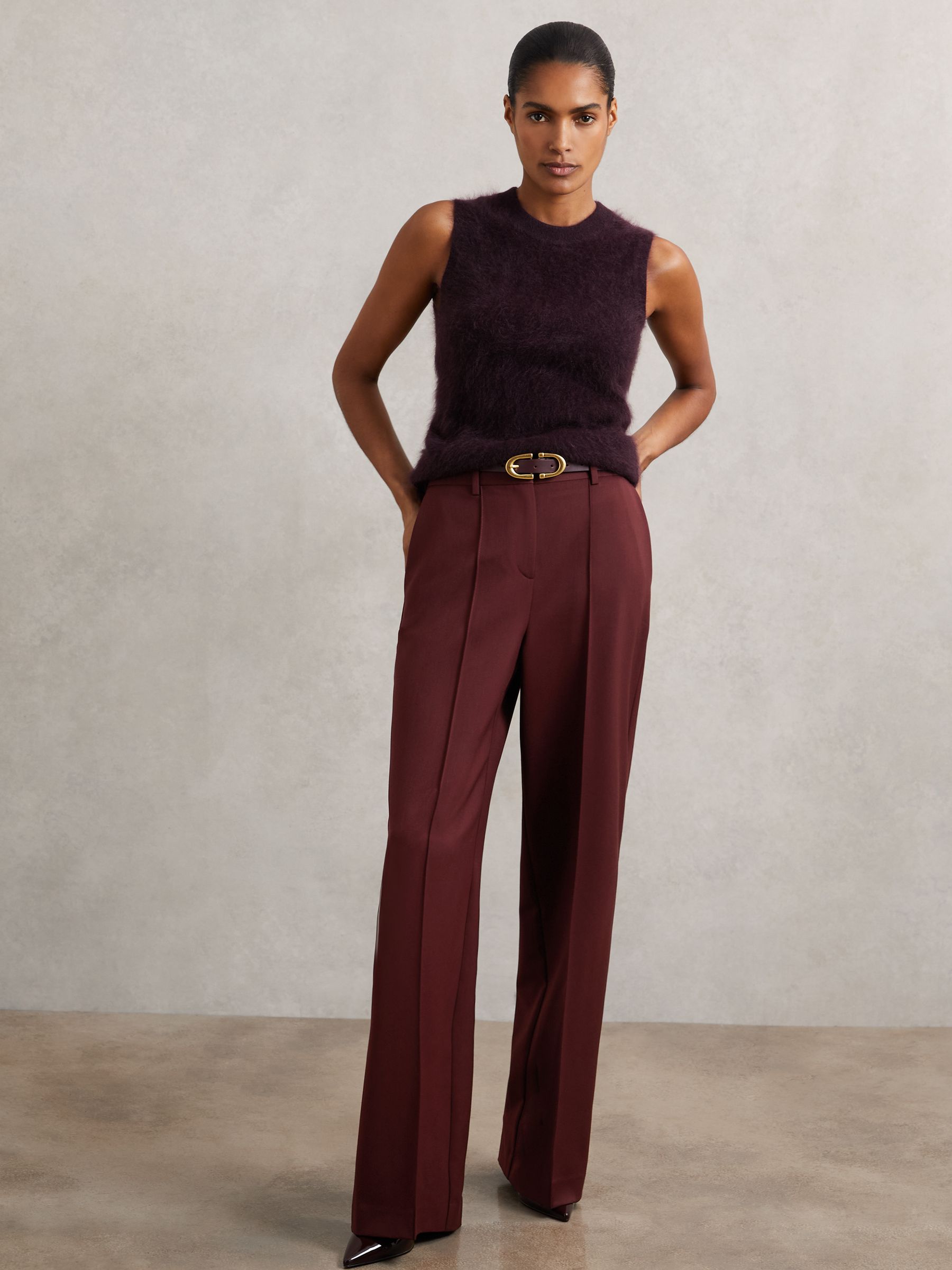 A woman wearing burgundy wide-leg trousers with front pintucks, side slip pockets, and back welt pockets, paired with a sleeveless top.