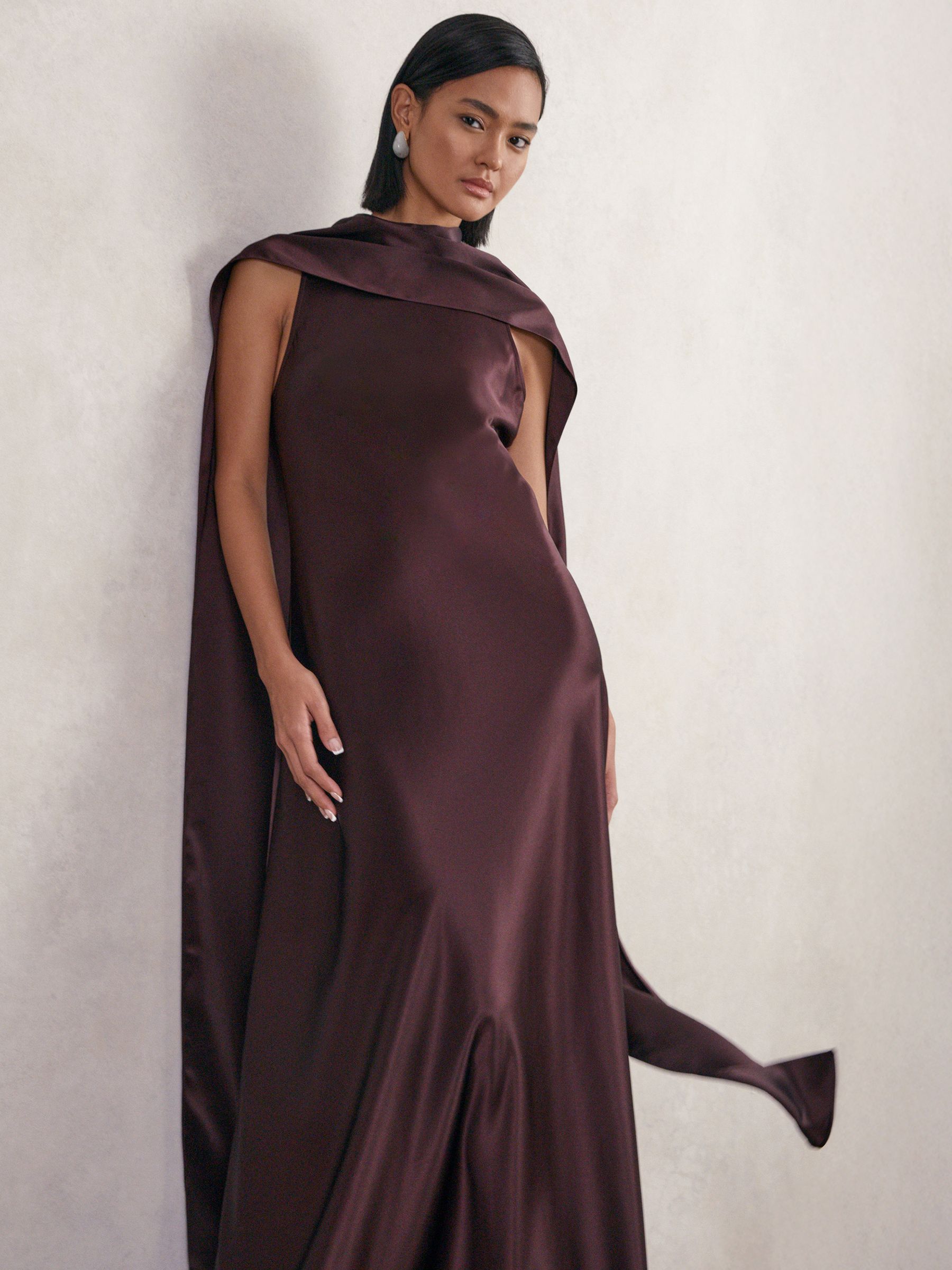 A woman wearing a burgundy satin maxi dress with a cape-style design, sleeveless cut, and a draped cowl neckline.