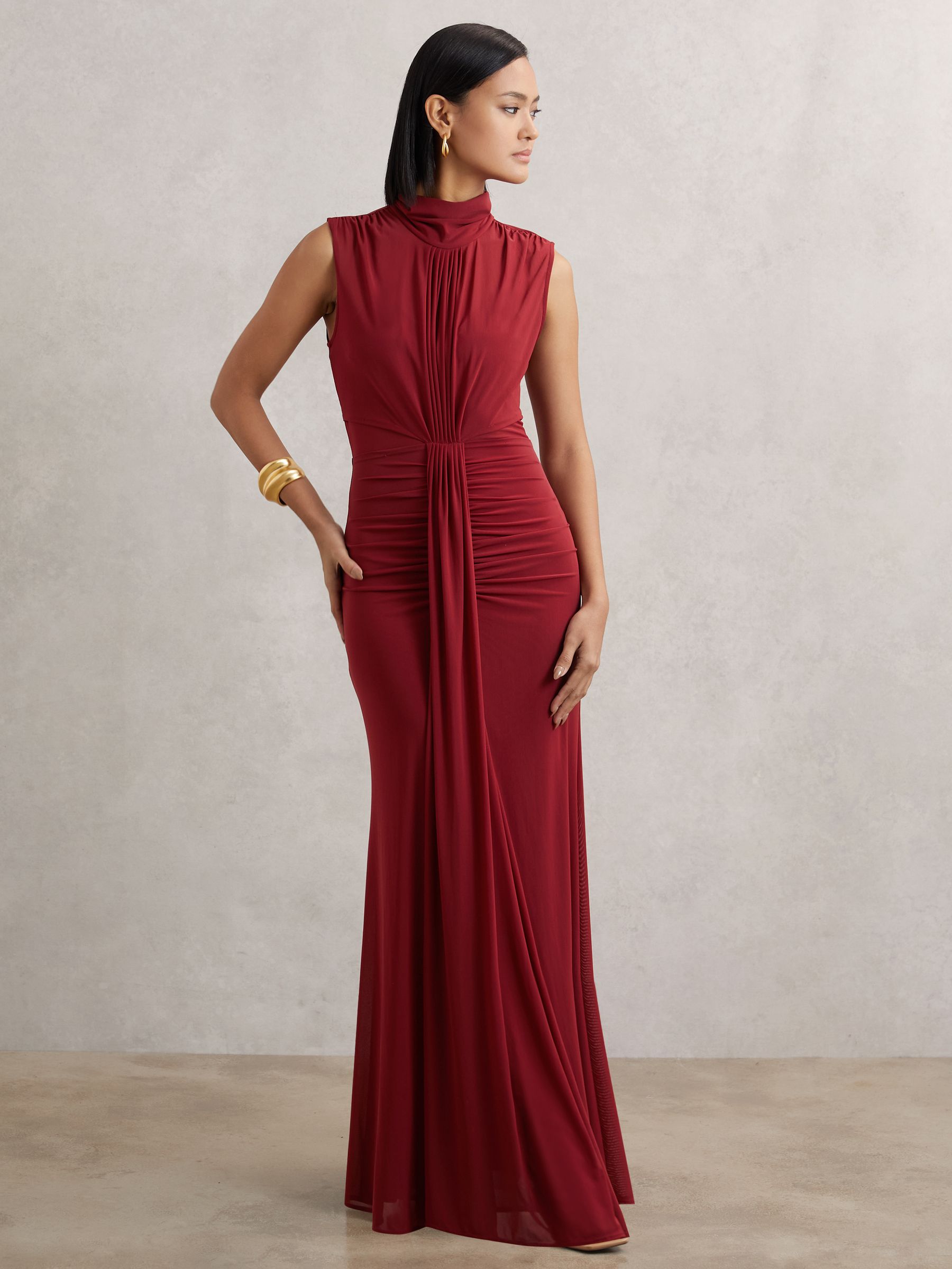 A woman wearing a sleeveless, dark red maxi dress with a high neckline, ruching at the center, sheer bodice detail, and a flared hem, standing against a plain background.