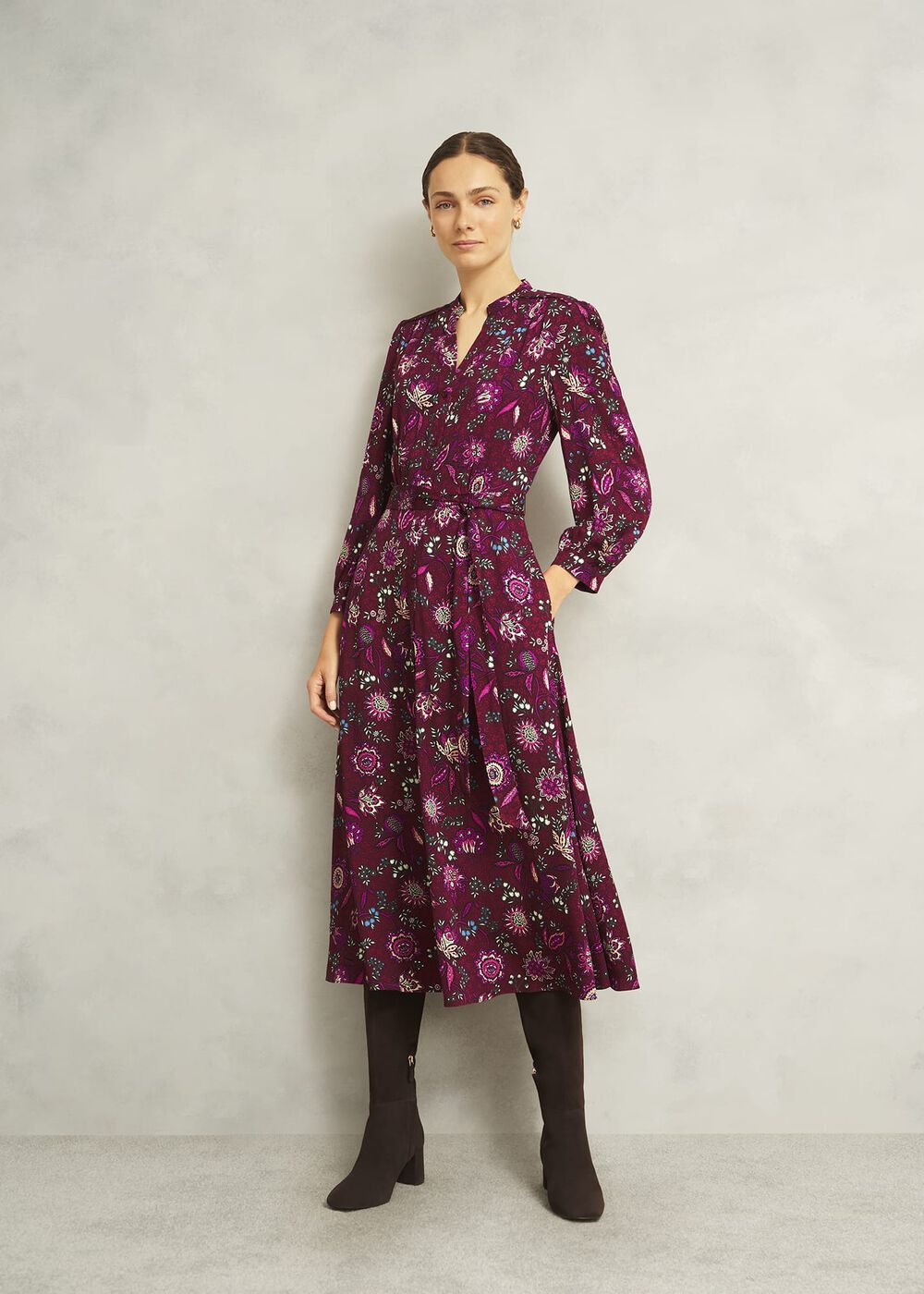 A woman wearing a magenta floral midi dress with a notch neckline, long sleeves, and a waist tie, paired with black knee-high boots.
