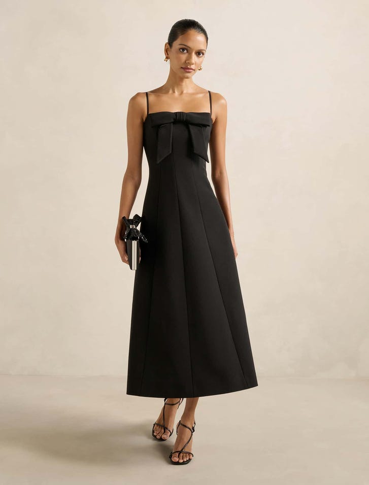 A woman wearing a black midi dress with a bow front detail, square neckline, and spaghetti straps, standing against a plain background.