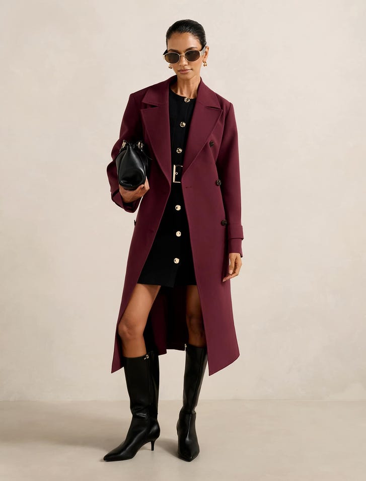 A woman wearing a deep burgundy Lydia Petite Crepe Trench Coat, black dress, black knee-high boots, sunglasses, and holding a black handbag.