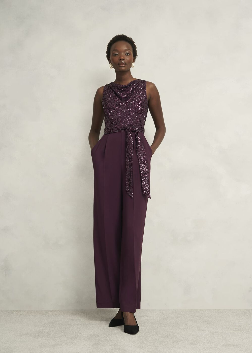 A woman wearing a purple sequined sleeveless jumpsuit with wide-leg trousers, a cowl neck, and a waist tie, standing against a plain background.