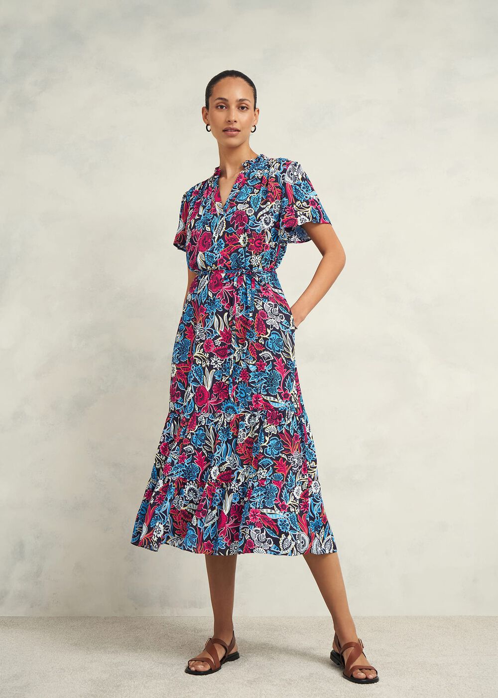 A woman wearing a midi-length floral dress with a mandarin collar, V-neckline, three-quarter sleeves, side pockets, and a tiered hem with a frill detail, in shades of pink, blue, white, and black.
