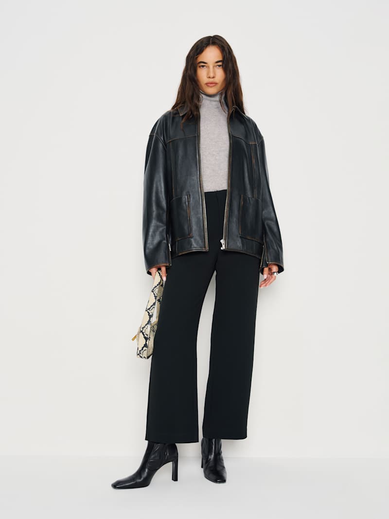 A woman wearing black high-rise cropped pants, paired with a grey turtleneck and black heeled boots, holding a patterned handbag.