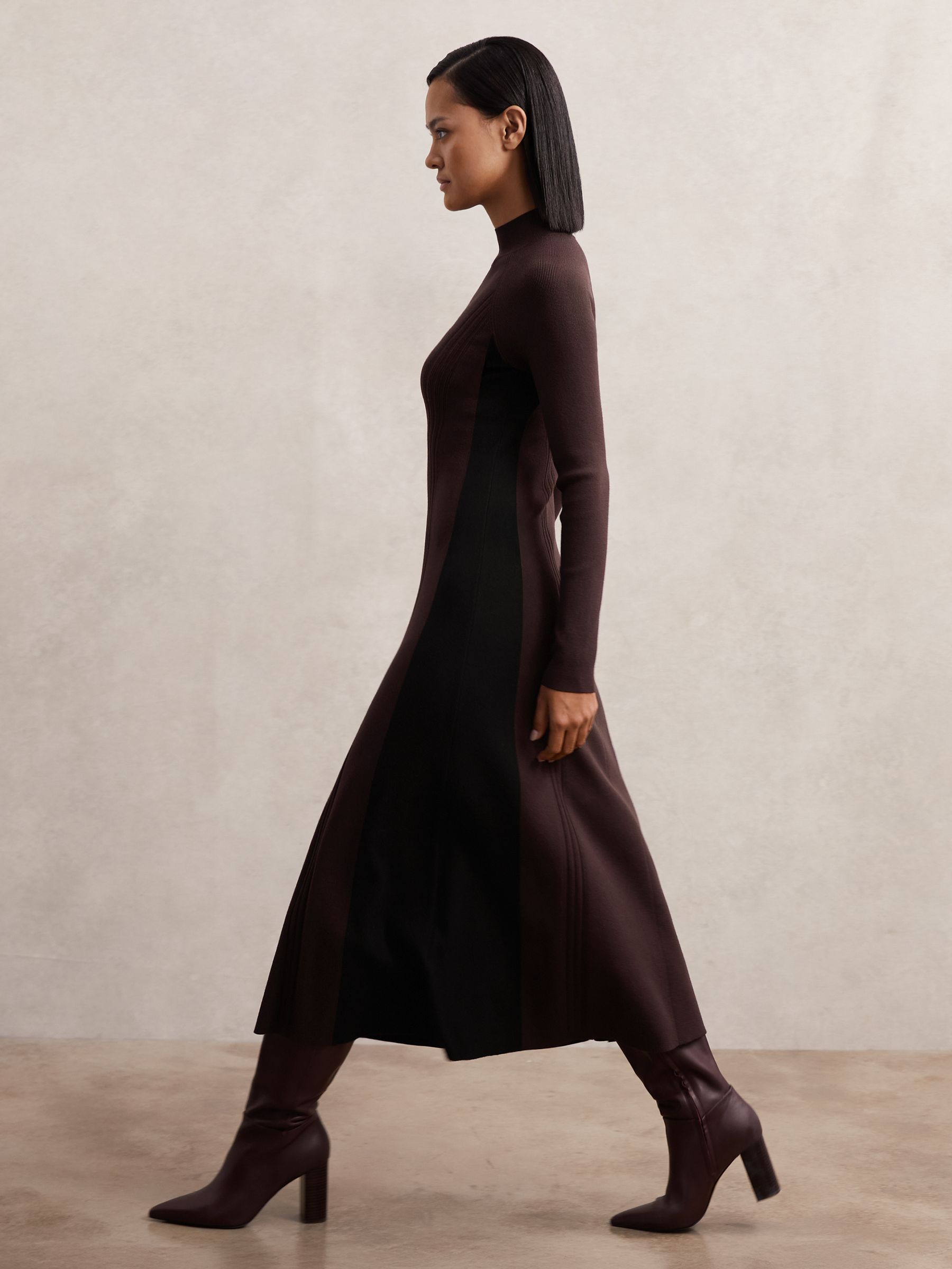 A woman wearing a knitted midi dress with contrast plum purple and black panels, ribbed details, and a fluted skirt, paired with high-heeled boots.