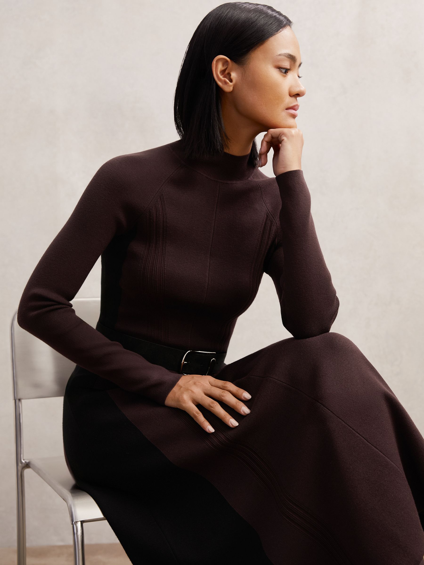 A woman wearing a plum purple and black contrast-panel midi dress with ribbed knitting, a mock neckline, and a fluted skirt.