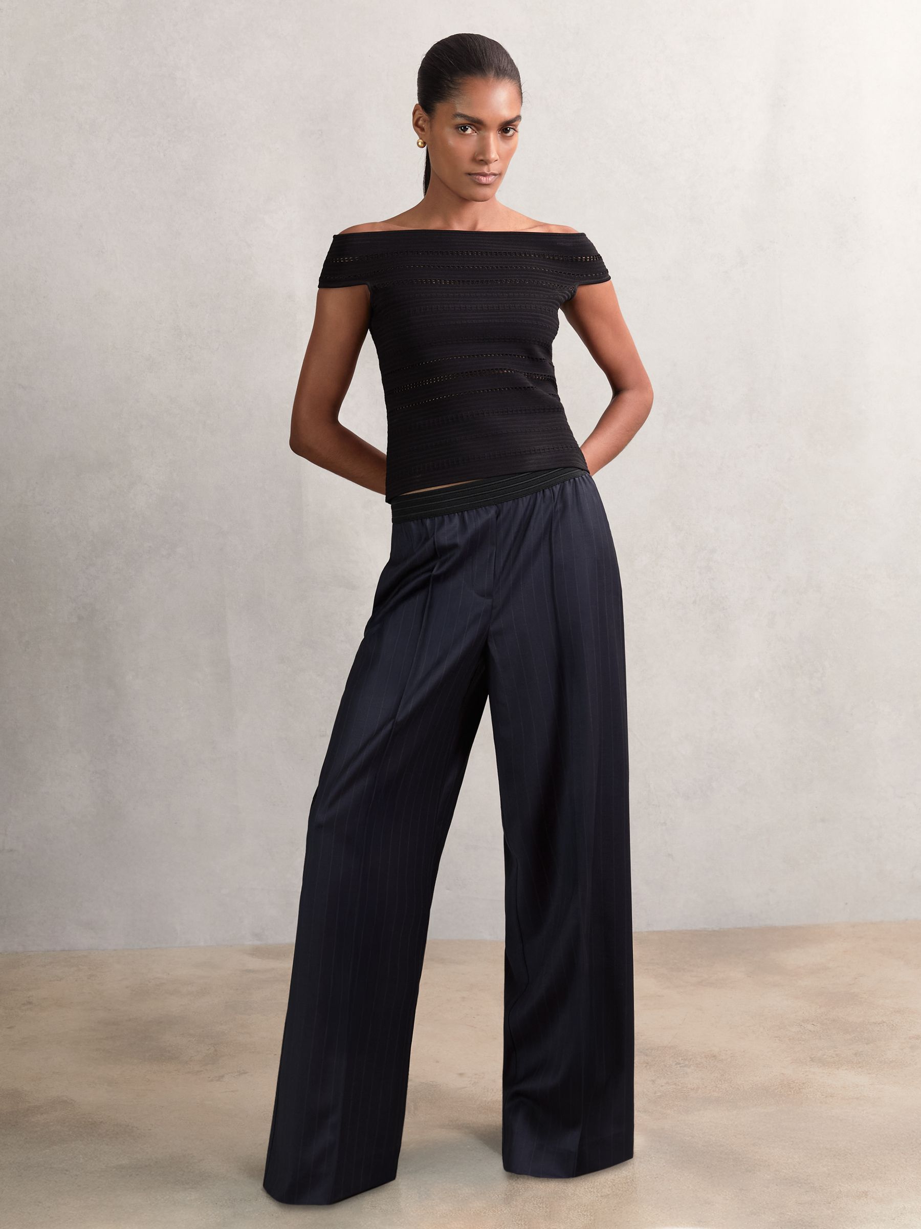 A woman wearing navy wide-leg trousers with pinstripes, an elastic waistband, and front pintucks, paired with a black off-shoulder top.