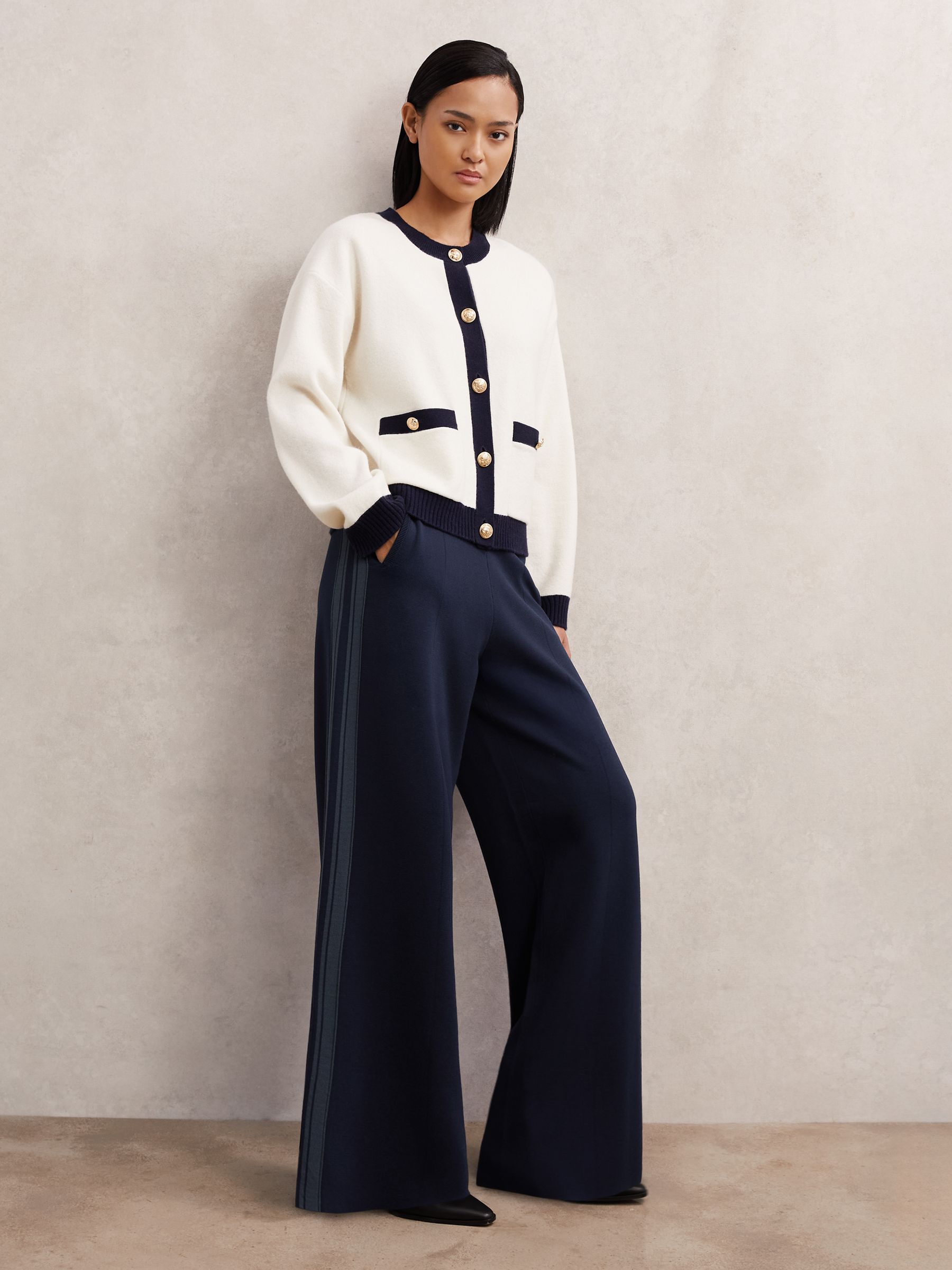 A woman wearing navy knitted wide-leg trousers with side stripes, an elastic waistband, and side pockets, paired with a white and navy cardigan.