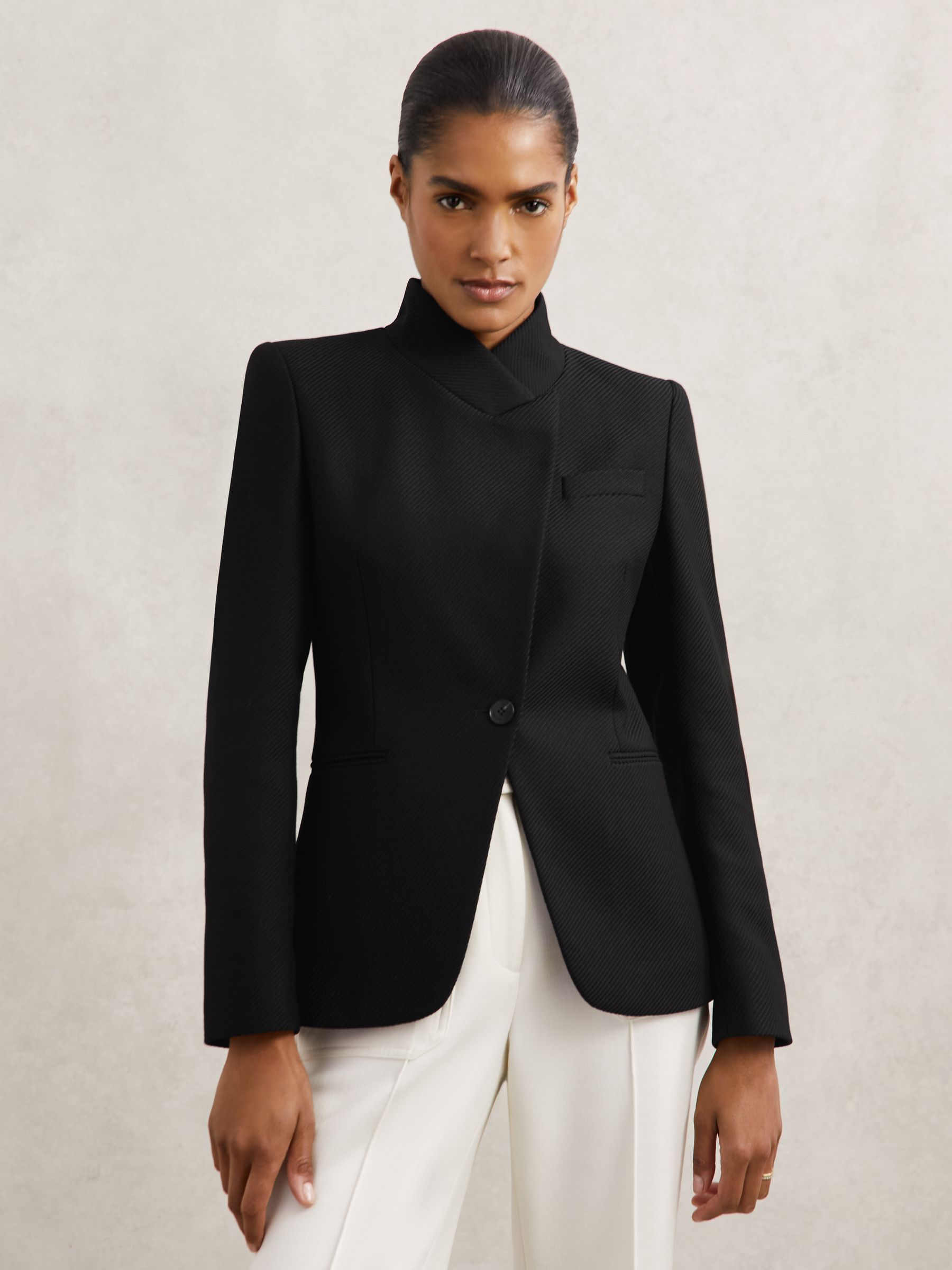 A woman wearing a black tailored blazer with a wrapover front, standing collar, and a single vent at the back.