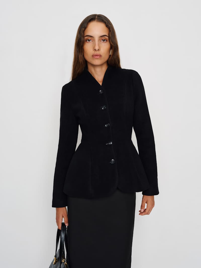 A woman wearing a black tailored jacket with a button front and curved hem, fitted at the waist, made from heavy weight fabric.