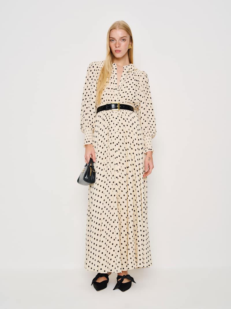 A woman wearing a beige maxi dress with black polka dots, puff sleeves, a button front, and a black belt, holding a small black handbag and wearing black shoes with bows.