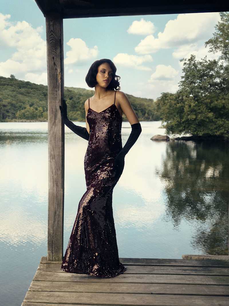 A woman wearing a dark, sequined, sleeveless maxi dress with a V-neckline and adjustable straps, standing on a wooden dock by a lake.