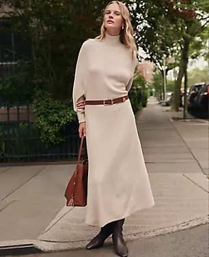 A woman wearing a beige ribbed turtleneck sweater dress with long raglan sleeves, drop shoulders, and a fabric tie belt, standing on a sidewalk with trees and parked cars in the background.
