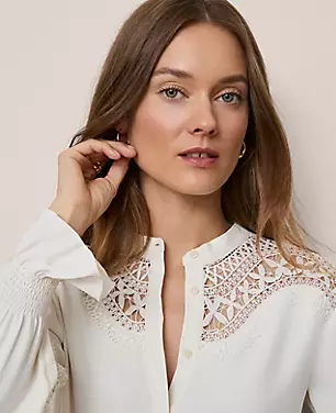 A woman wearing a white blouse with embroidered lace details on the shoulders, stand collar, hidden buttons, and elasticized cuffs.