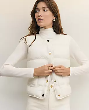 A woman wearing a white quilted vest with a crewneck, sleeveless design, and snap buttons, standing against a plain background.