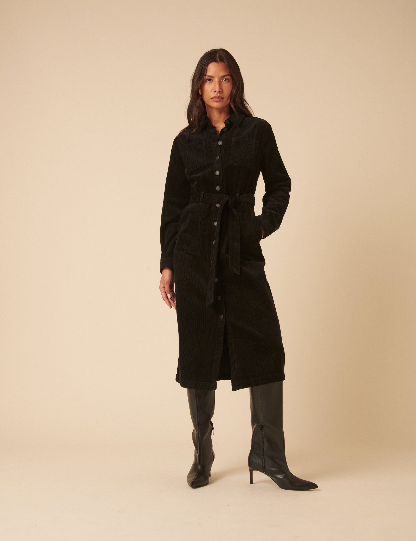 A woman wearing a black midi shirt dress with long sleeves, a tie belt, and side pockets, standing against a plain background.