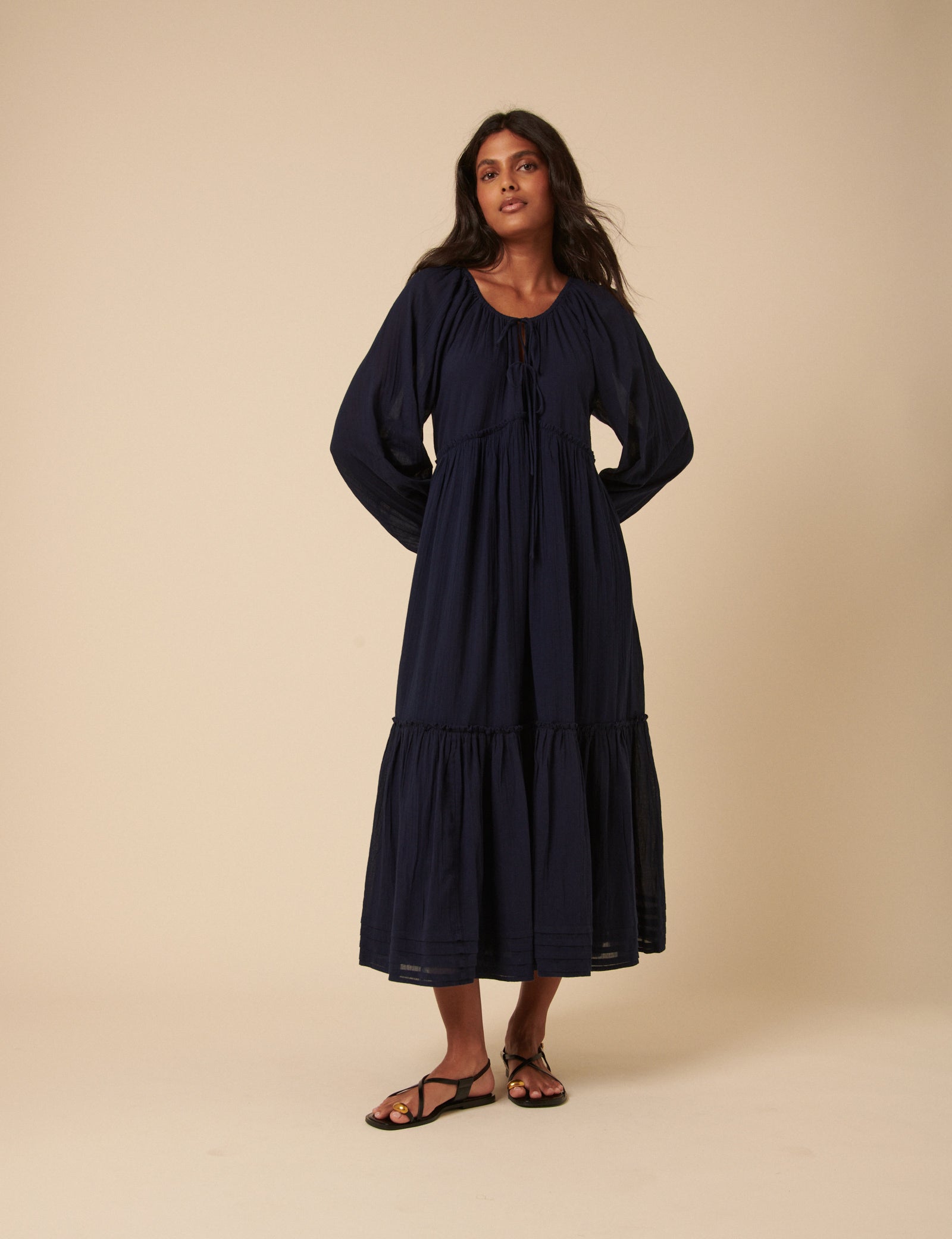 A woman wearing a navy midi dress with a scoop neckline, long sleeves, and a tiered skirt with a front tie detail.