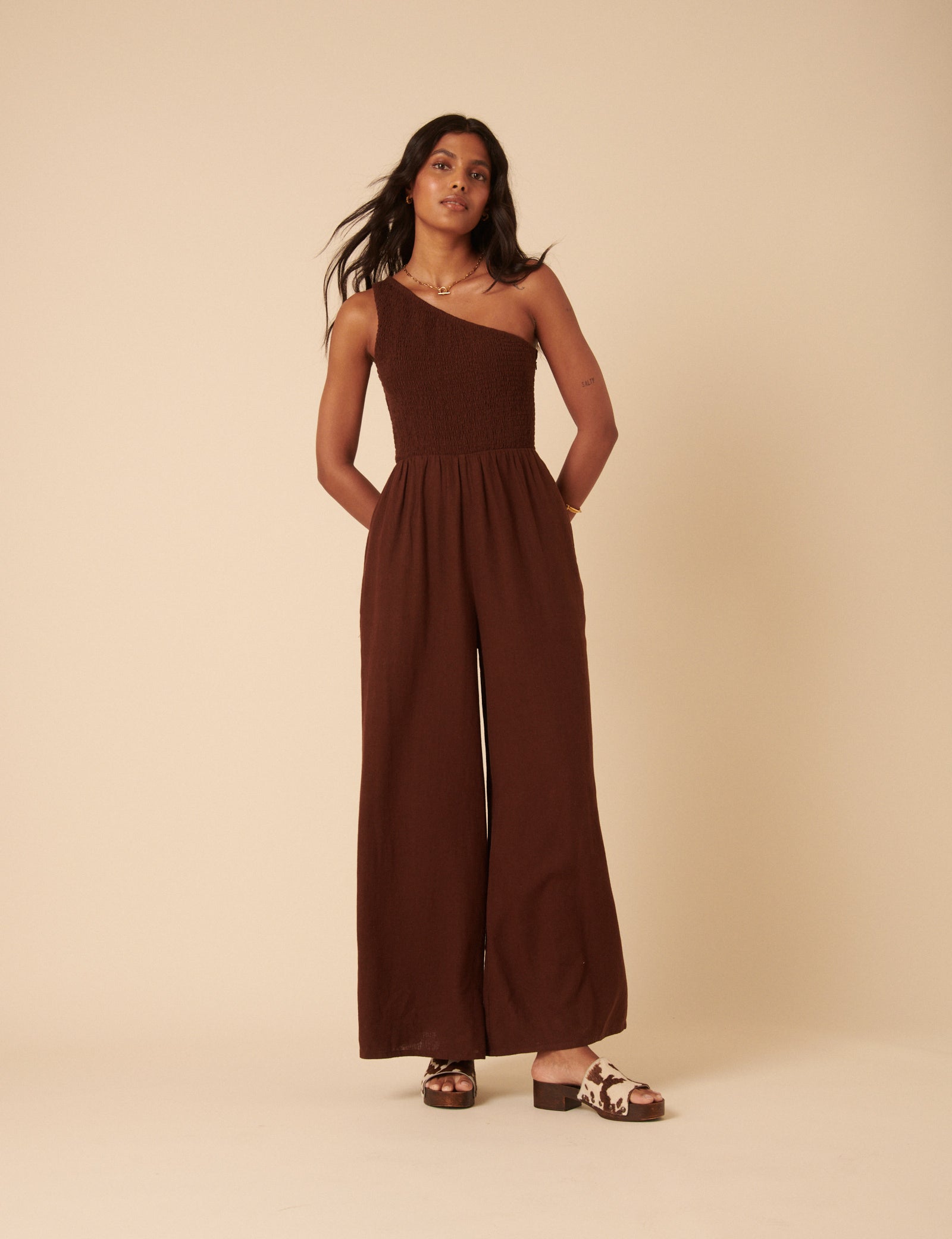 A woman wearing a brown one-shoulder jumpsuit with a ruched bodice and wide-leg pants, standing against a plain background.
