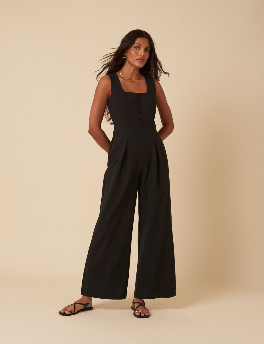 A woman wearing a black jumpsuit with a scoop neckline, button details on the front, and wide-leg pants.