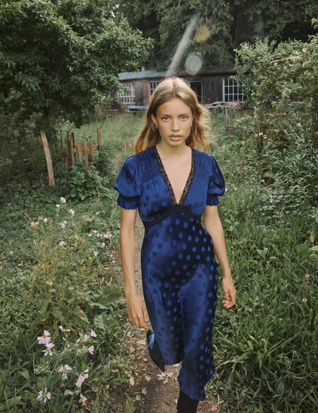 A woman wearing a navy blue midi dress with polka dots, V-neckline, short puff sleeves, and a relaxed skirt, standing outdoors in a garden.