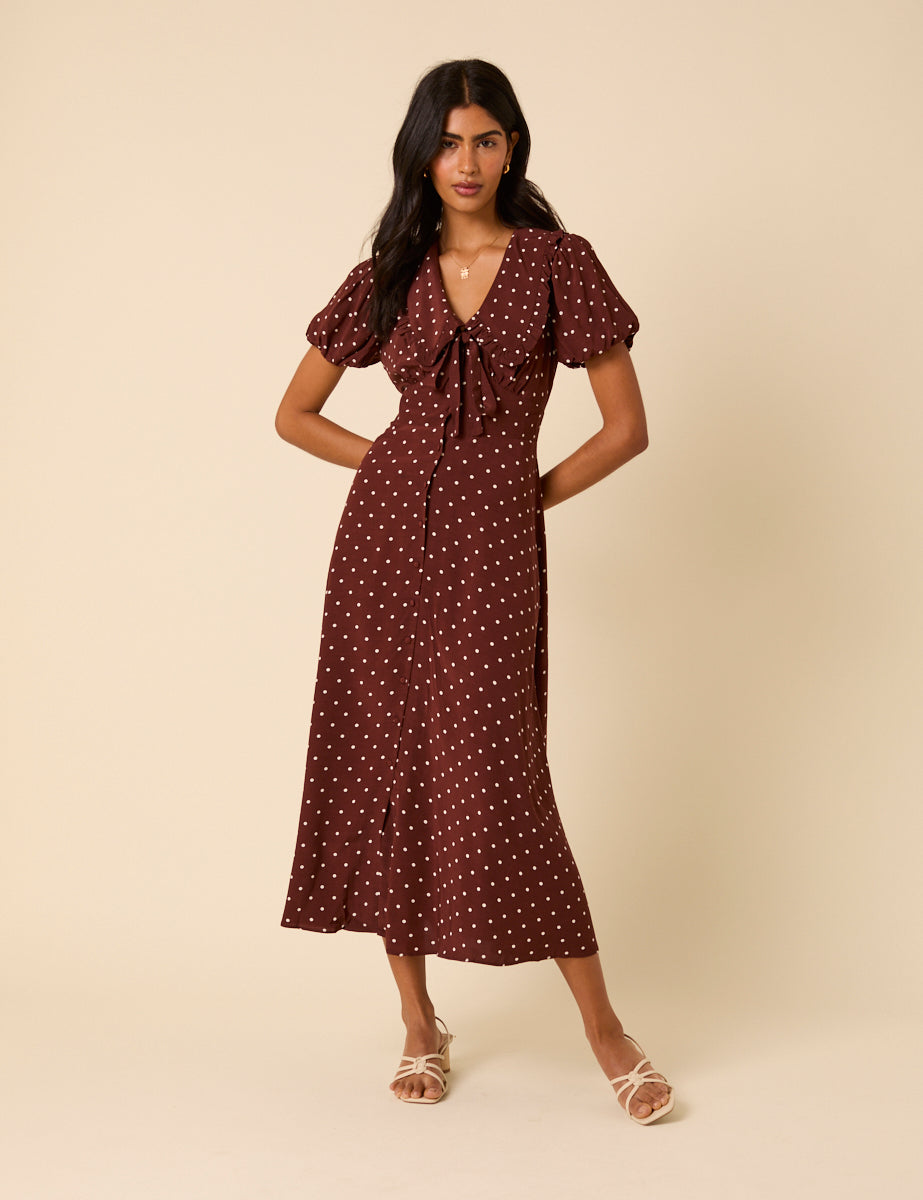 A woman wearing a brown polka dot midi dress with puff sleeves, V-neckline, and a front tie, standing against a beige background.