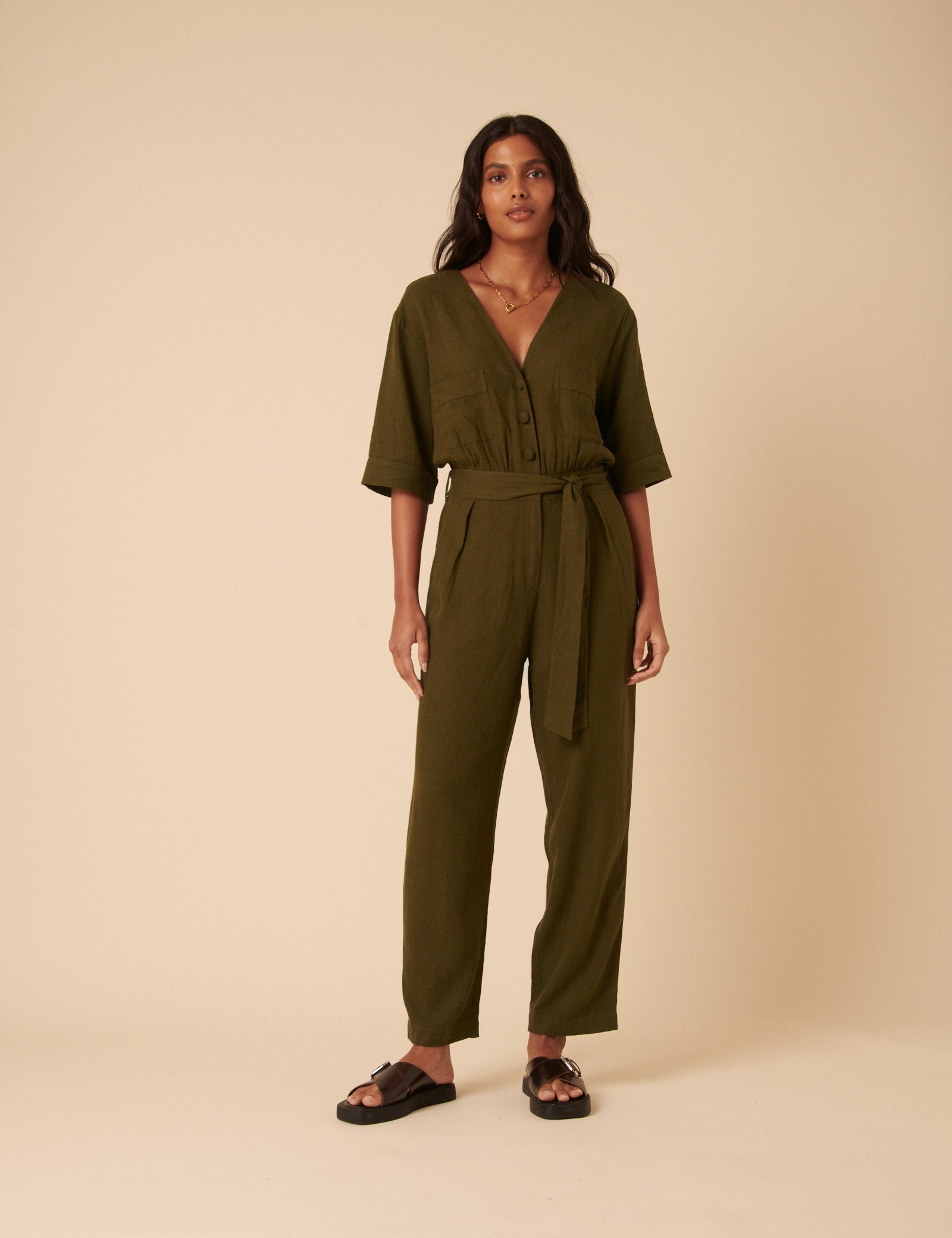 A woman wearing a khaki green linen-blend jumpsuit with a V-neckline, button-front, front pockets, and a waist tie.