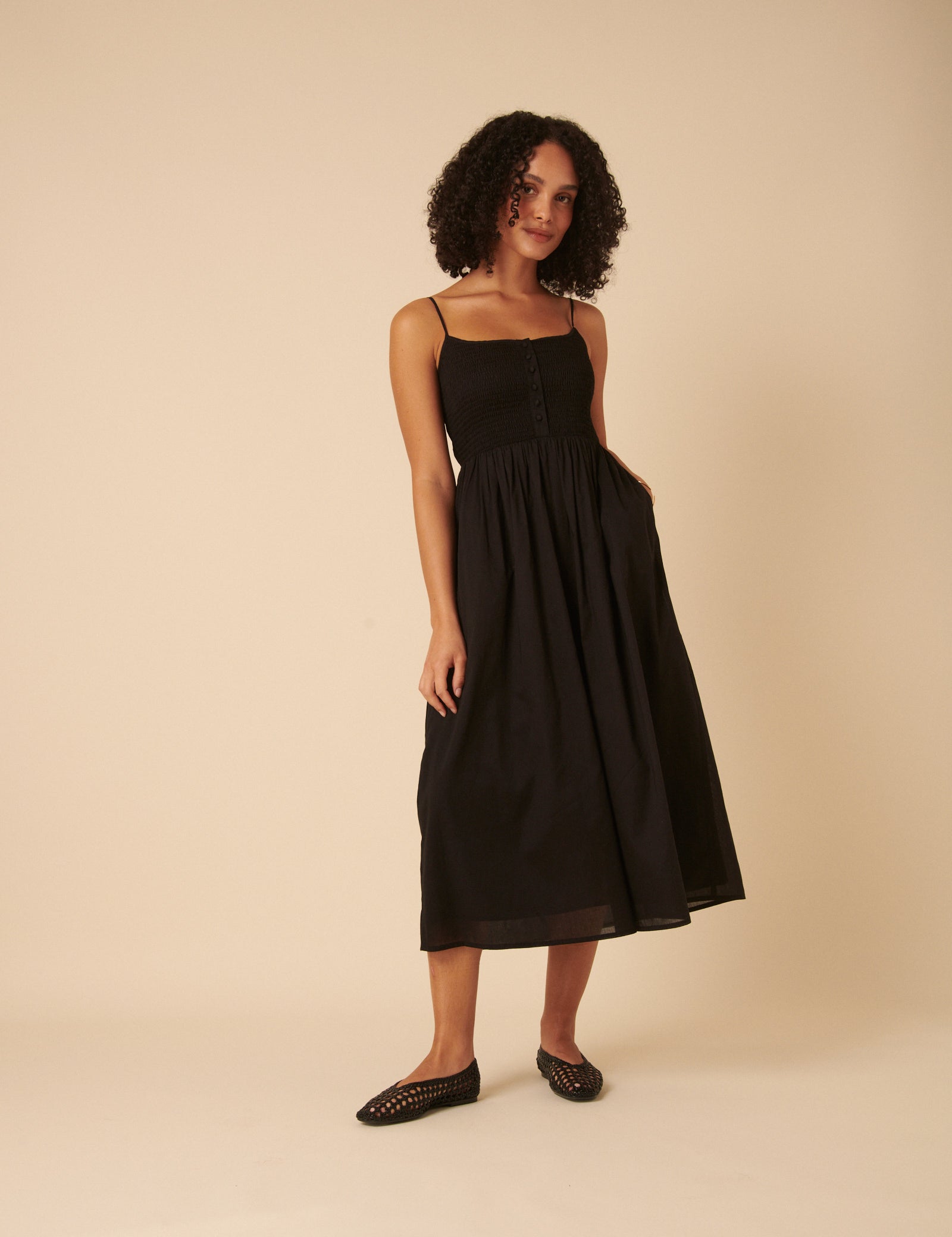 A woman wearing a black midi dress with spaghetti straps, a shirred bodice, and a full skirt, standing against a plain background.