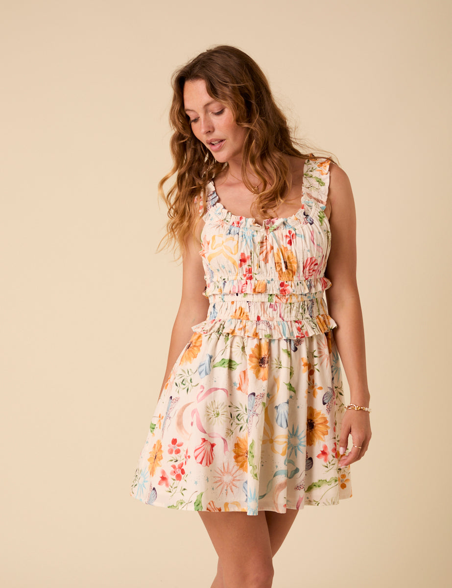 A woman wearing a white mini dress with a colorful floral and summer motifs print, square neckline with ruffle trims, and sleeveless design.