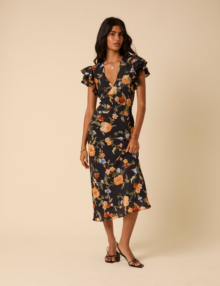 A woman wearing a black floral midi dress with ruffle sleeves and a V-neckline, standing against a beige background.