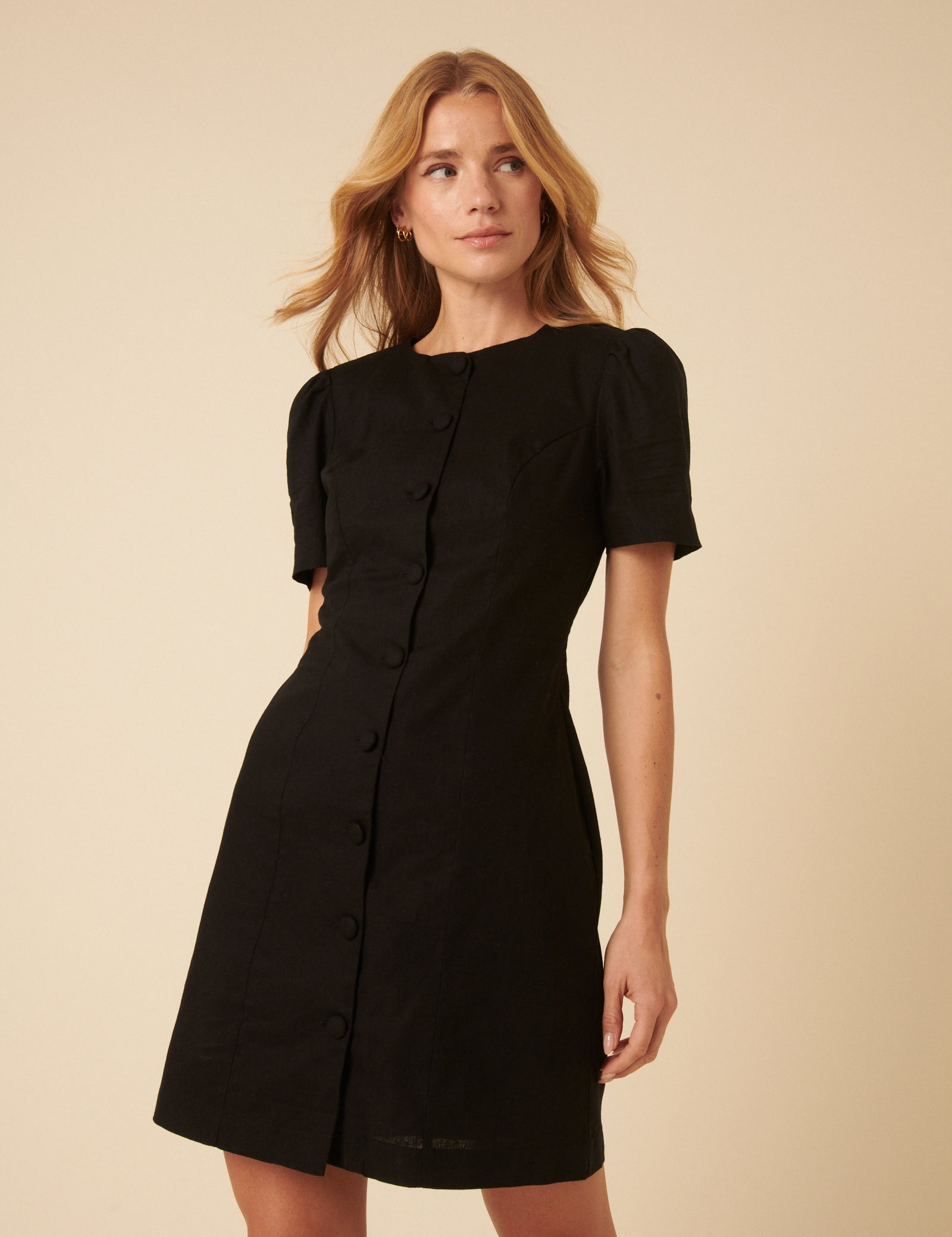 A woman wearing a black mini dress with a round neckline, short sleeves, and covered buttons down the front, with an A-line silhouette.