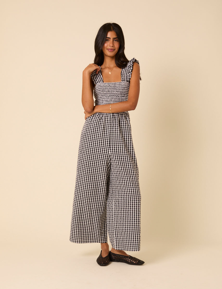 A woman wearing a black and white gingham jumpsuit with a square neckline, shirred bodice, wide-leg pants, and ruffled shoulder straps.