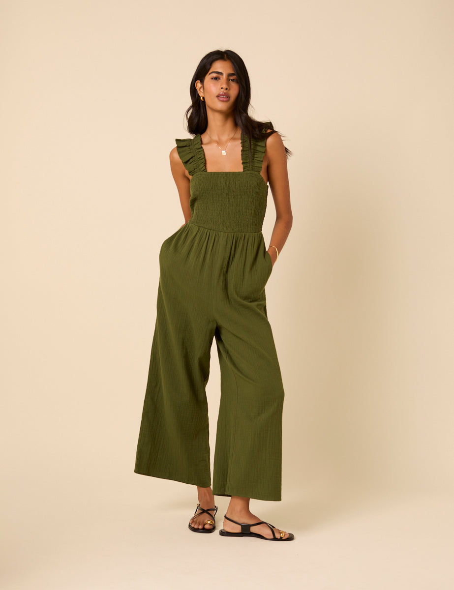 A woman wearing a khaki green cropped jumpsuit with ruffled straps, square neckline, and wide-leg pants, standing against a beige background.