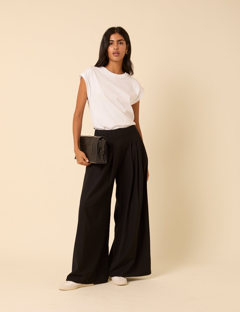 A woman wearing high-waisted black pleated wide leg trousers, paired with a white top and holding a black handbag.