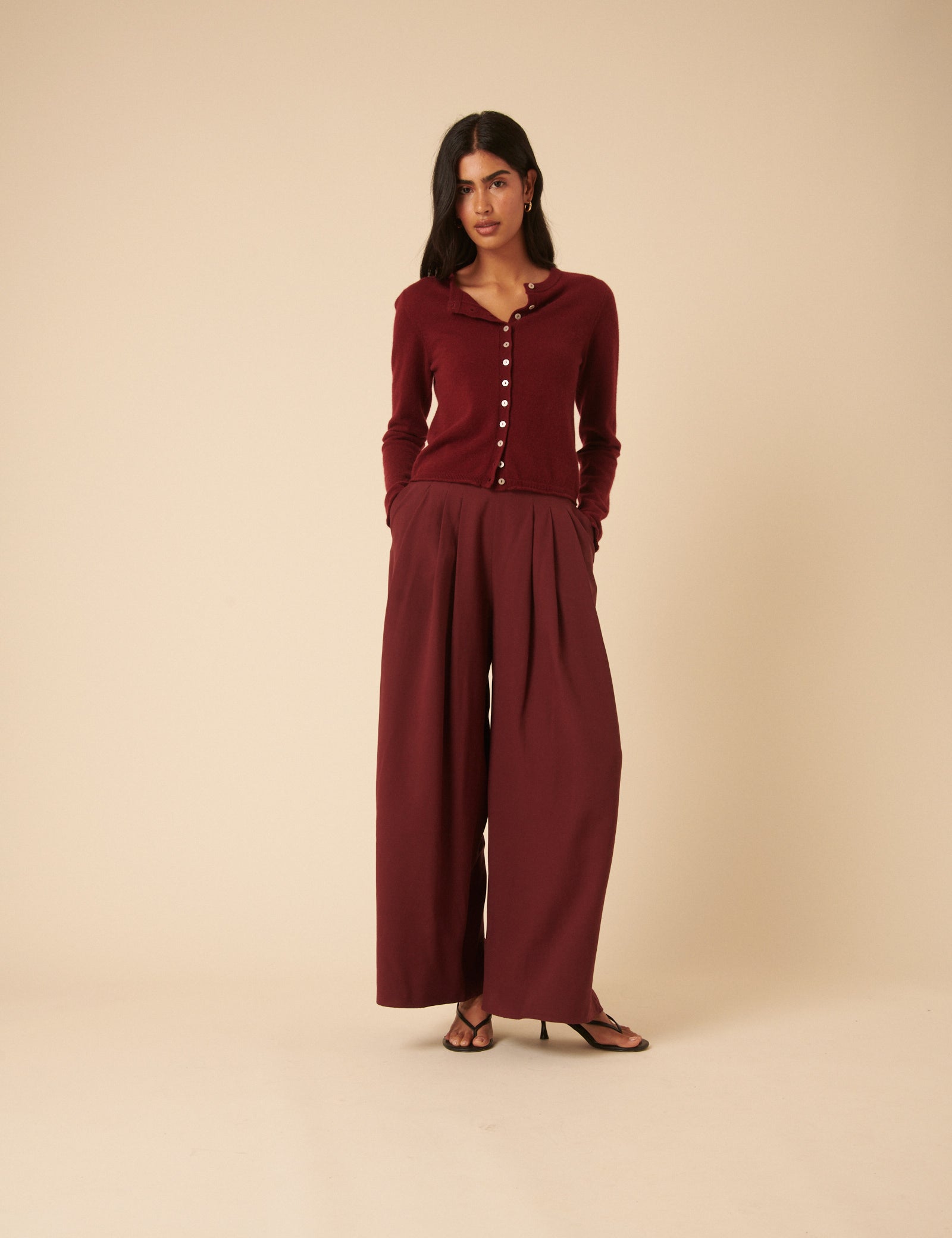 A woman wearing burgundy high-waisted pleated wide-leg trousers with side pockets, paired with a matching burgundy top.
