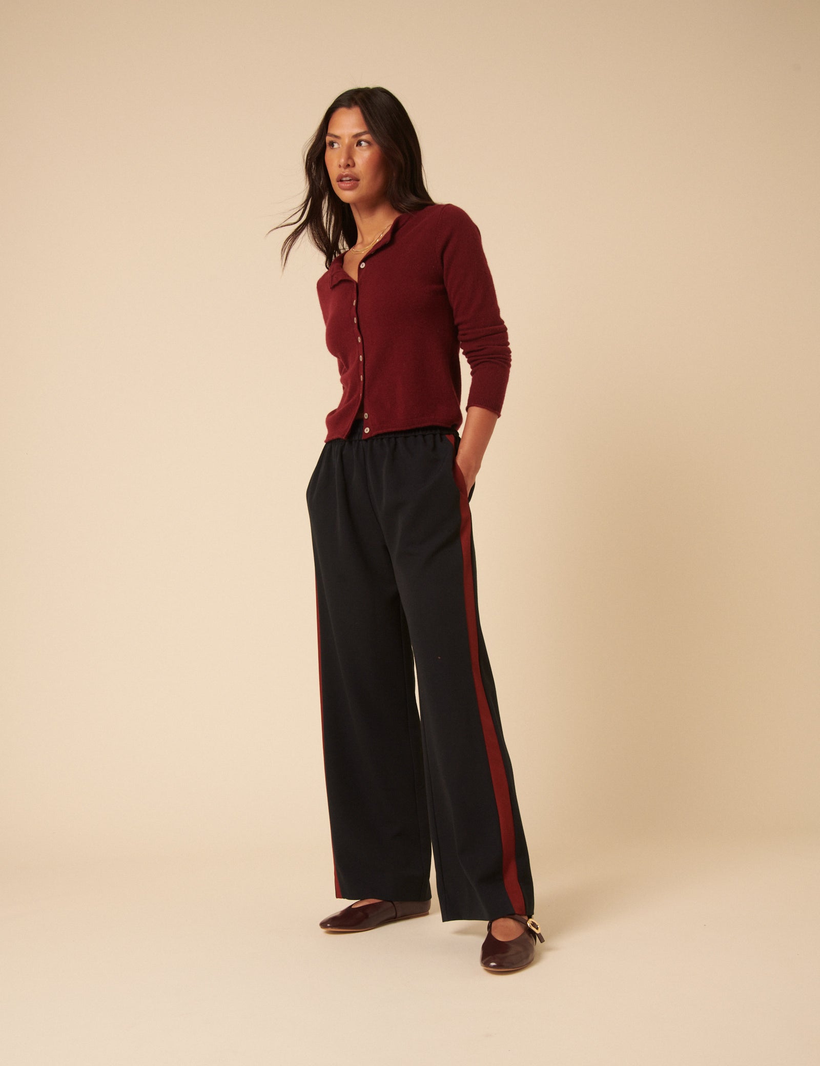 A woman wearing navy high-waisted trousers with red side stripes, paired with a burgundy long-sleeve top, standing against a beige background.