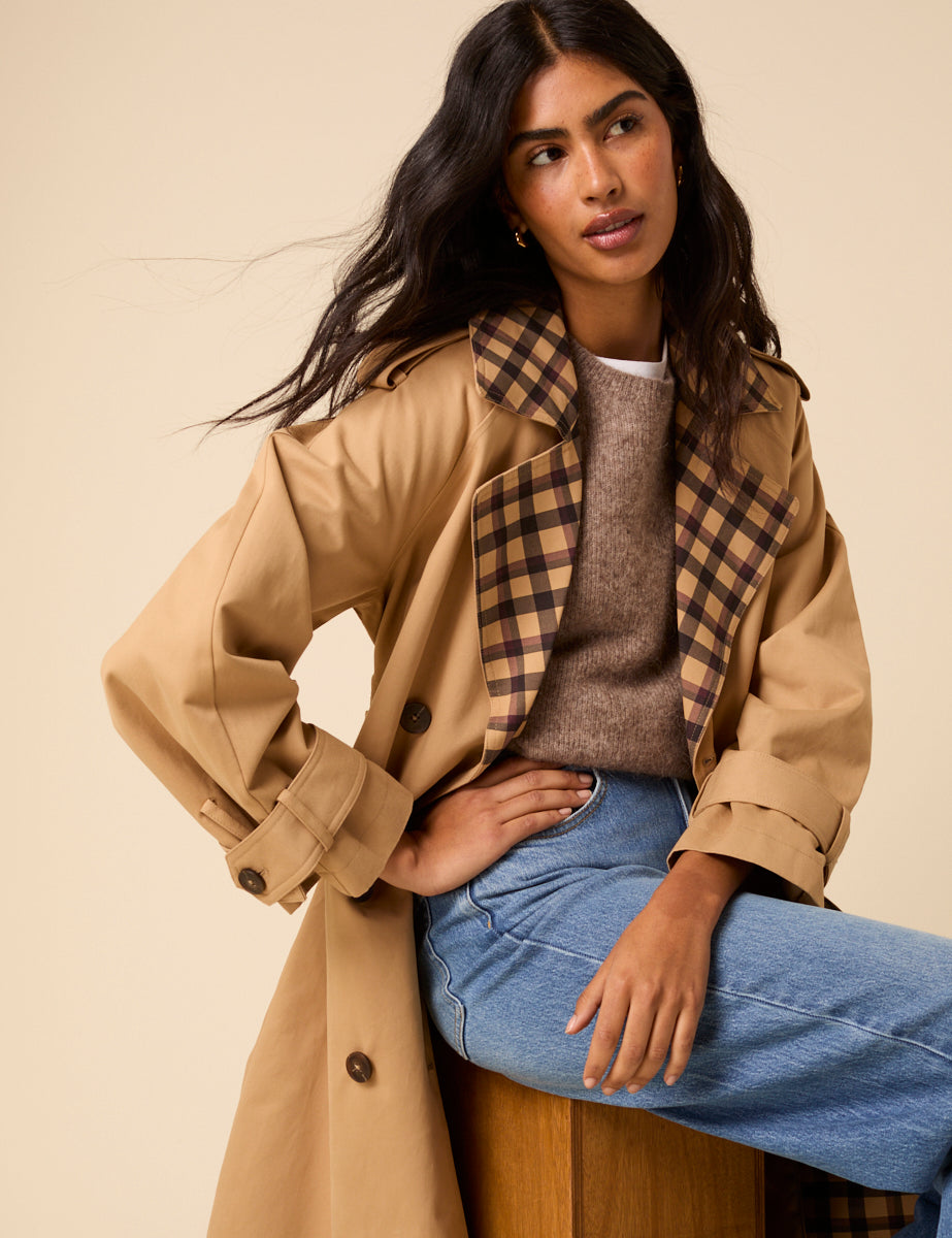 A woman wearing a beige trench coat with a camel check pattern on the collar and trim, featuring double-breasted buttons, a waist tie, epaulettes, and a collared neckline.