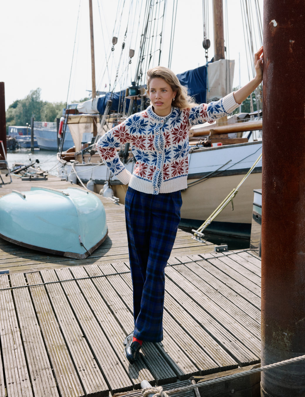 A woman standing on a wooden dock near boats, wearing navy check tailored trousers and a patterned sweater.