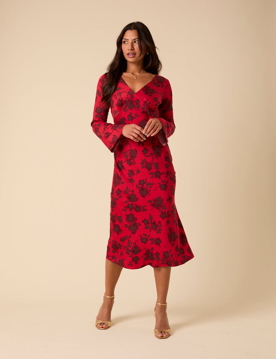 A woman wearing a red floral midi dress with a V-neckline and long fluted sleeves, standing against a beige background.