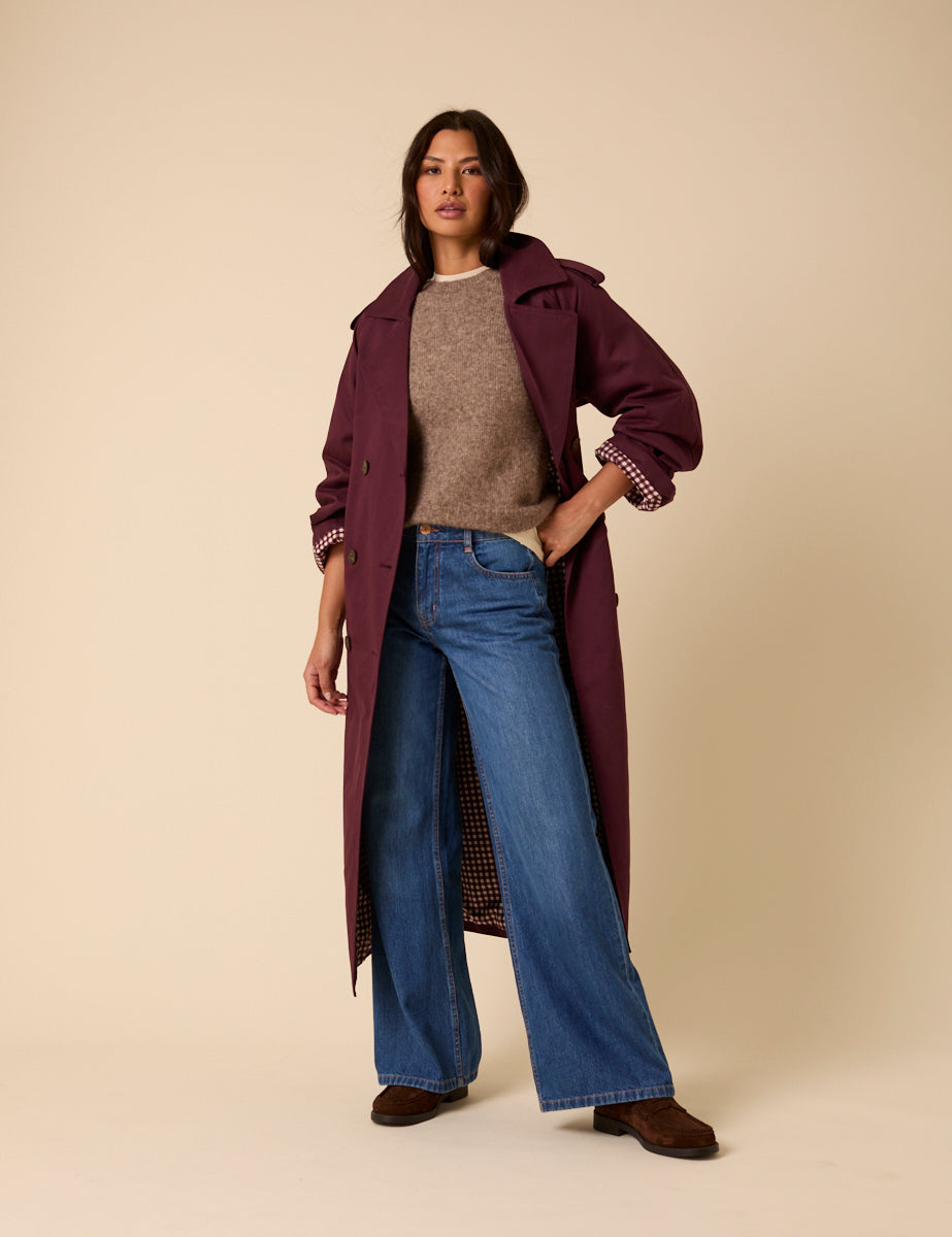 A woman wearing a burgundy double-breasted trench coat with a tie belt, epaulettes, and a collared neckline.