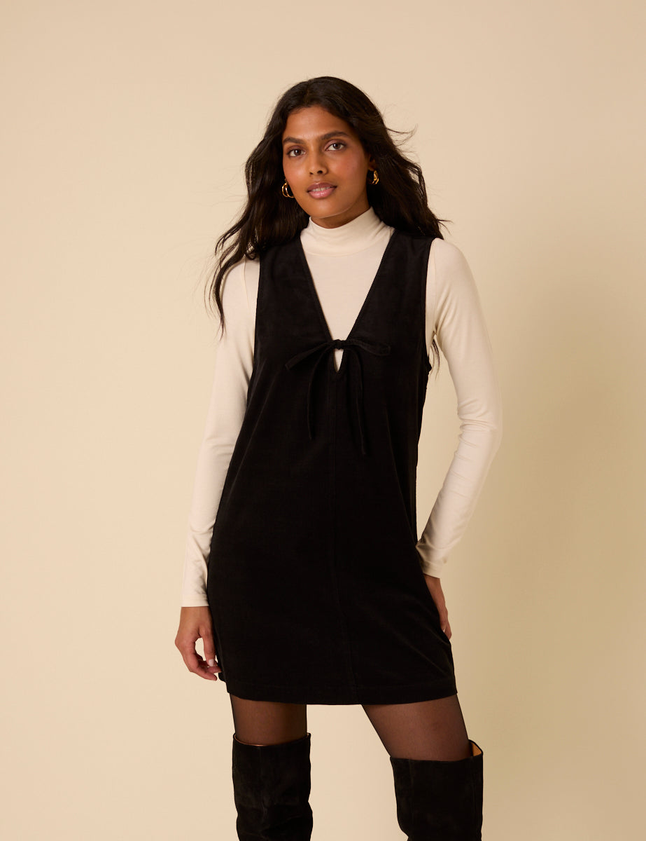 A woman wearing a black sleeveless mini dress with a V-neckline and tie detail, paired with a cream turtleneck top underneath.