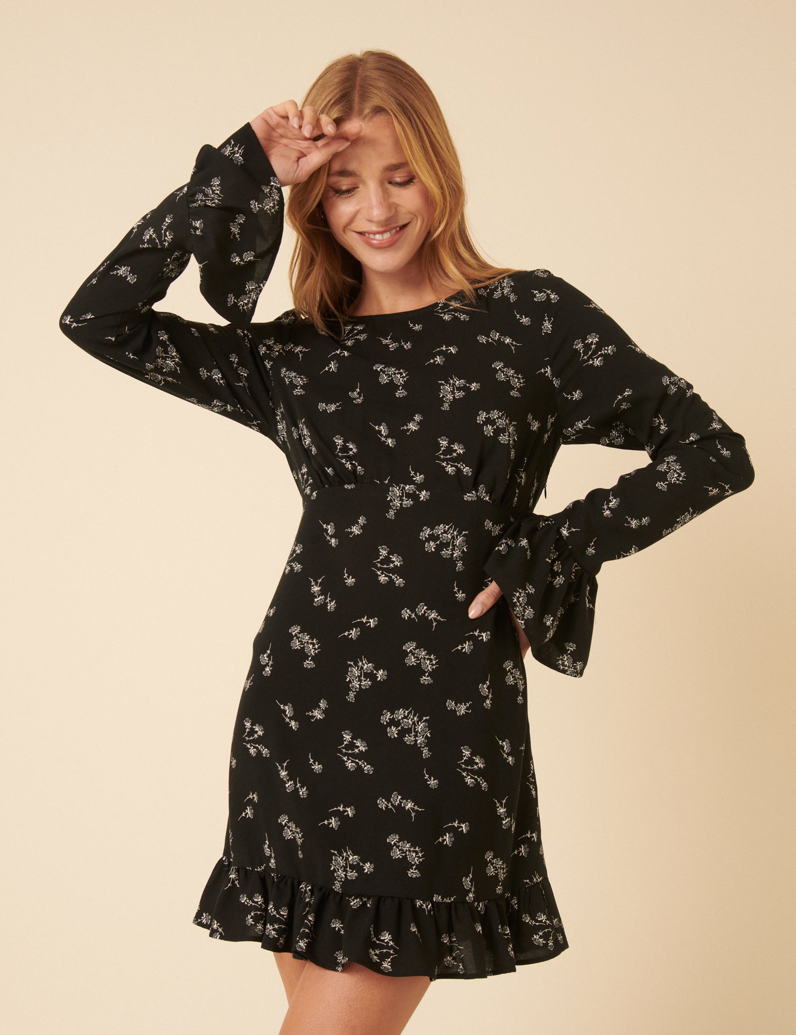 A woman wearing a black mini dress with white floral print, long fluted sleeves with ruffle cuffs, and a ruffled hem.