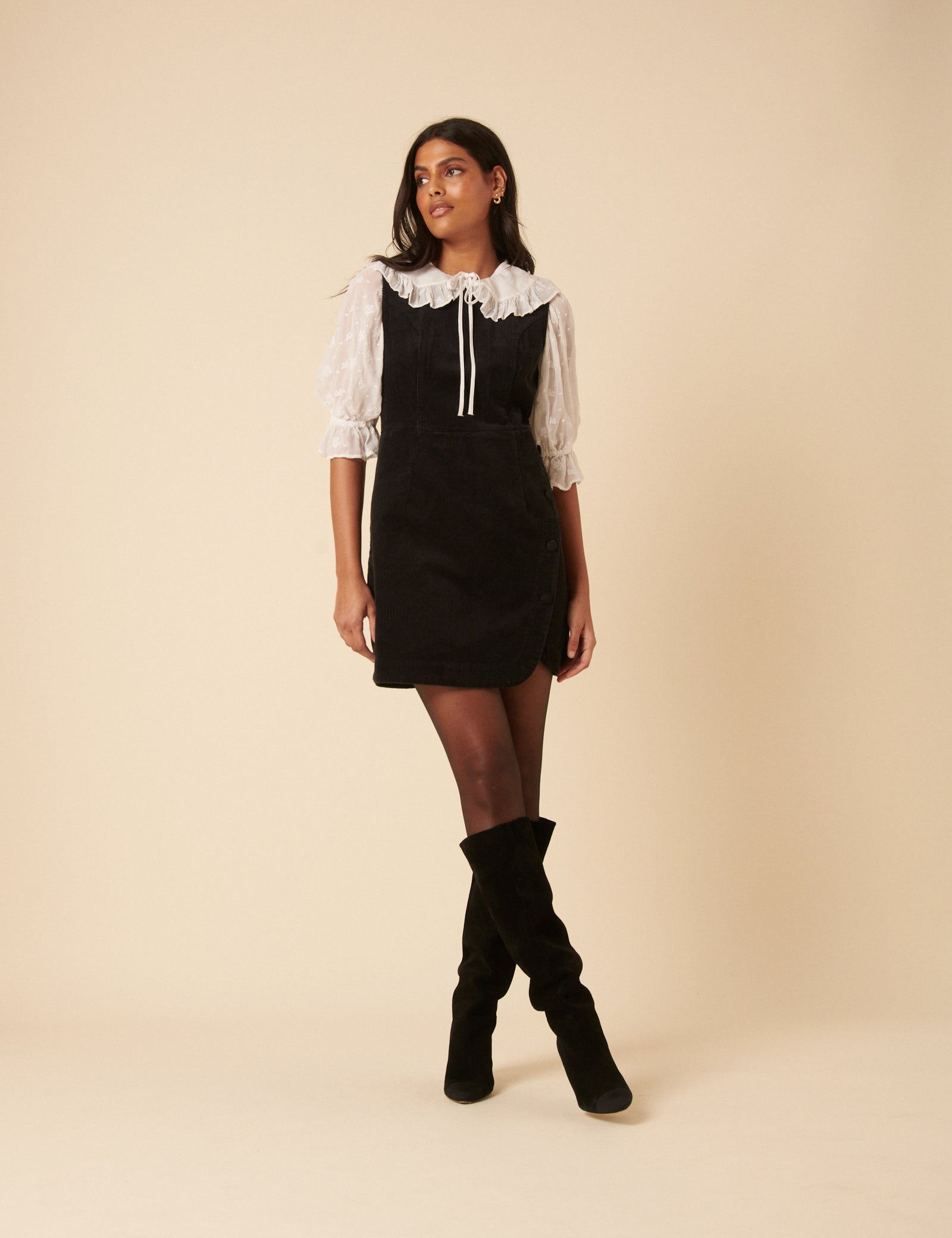 A woman wearing a black pinafore mini dress with a white ruffled collar and puffed sleeves, paired with black knee-high boots.