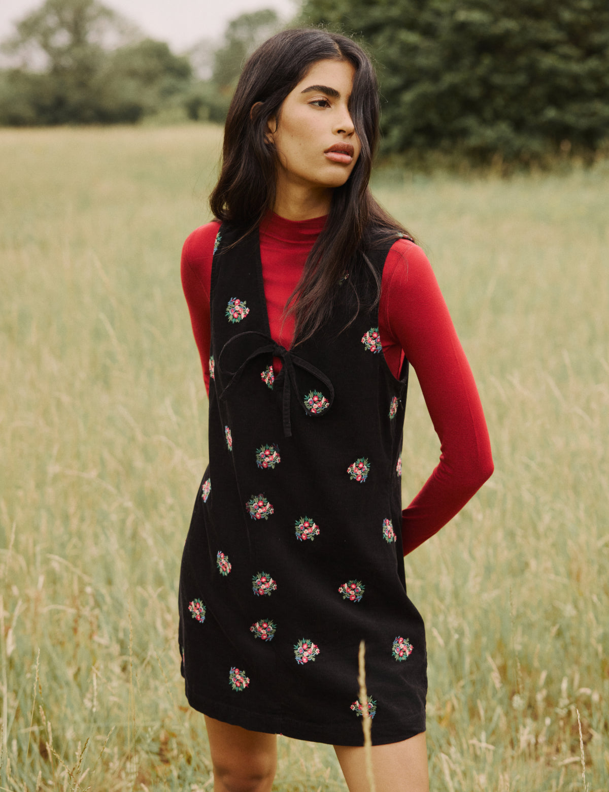 A woman wearing a black floral embroidered cord Nila Pinafore mini dress with a V-neckline and tie detail, standing outdoors in a grassy field.