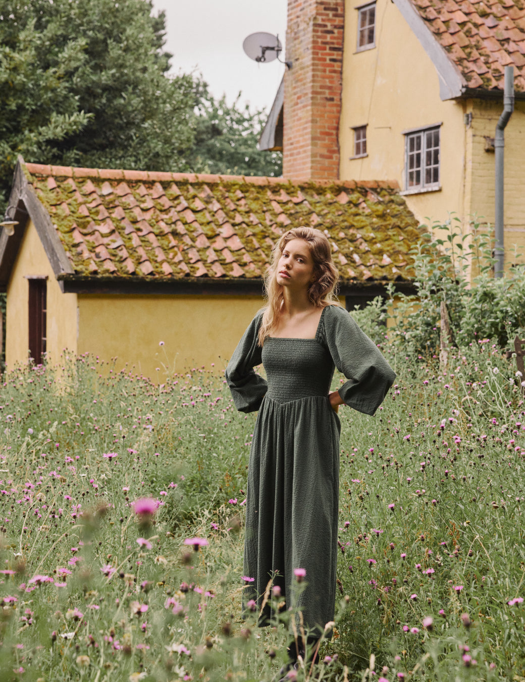A woman wearing a khaki long sleeve midi dress with a square neckline and shirred details, standing in a garden with yellow houses in the background.