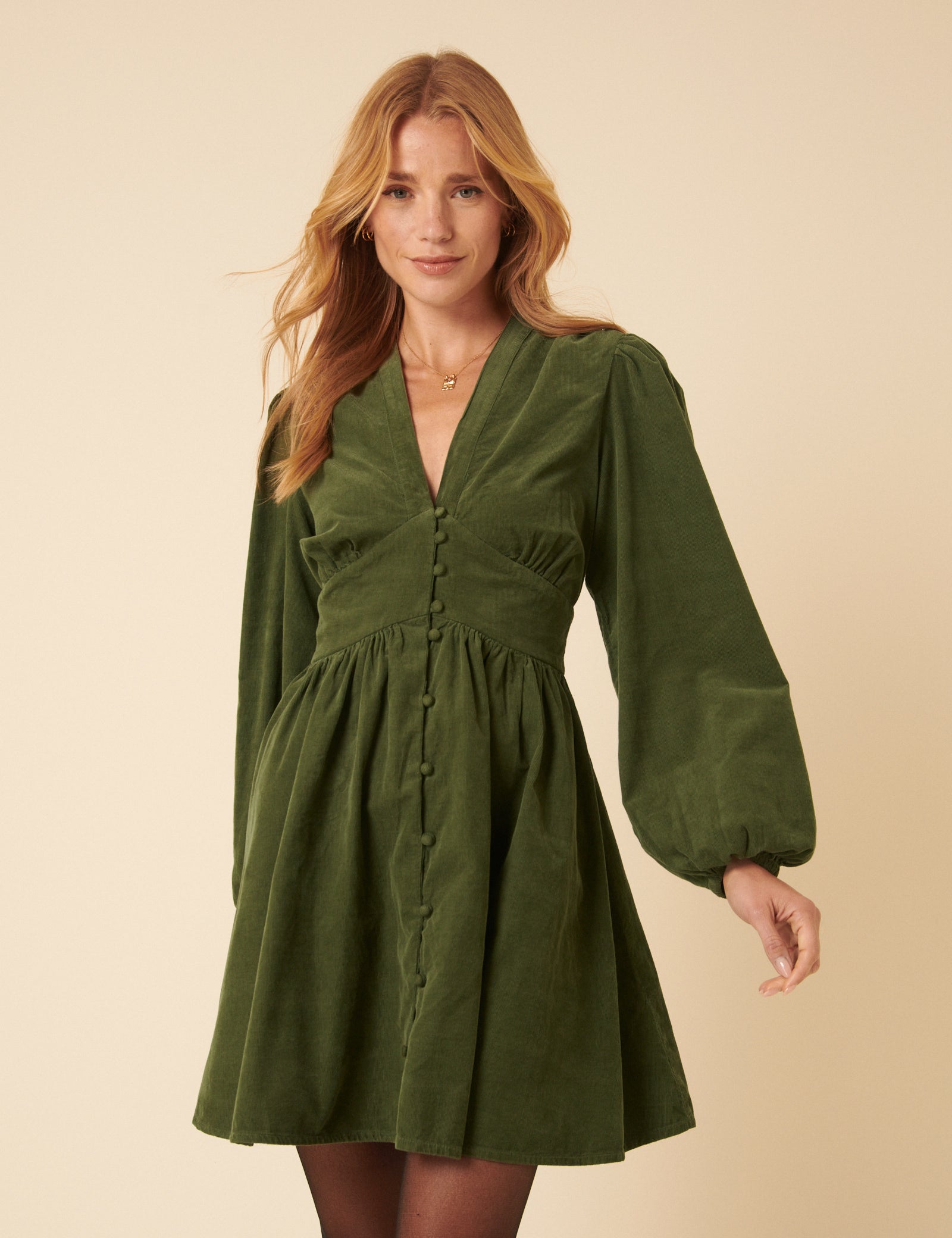 A woman wearing an olive green corduroy mini dress with a V-neckline, long sleeves, and buttons down the front, with side pockets and a flowy skirt.