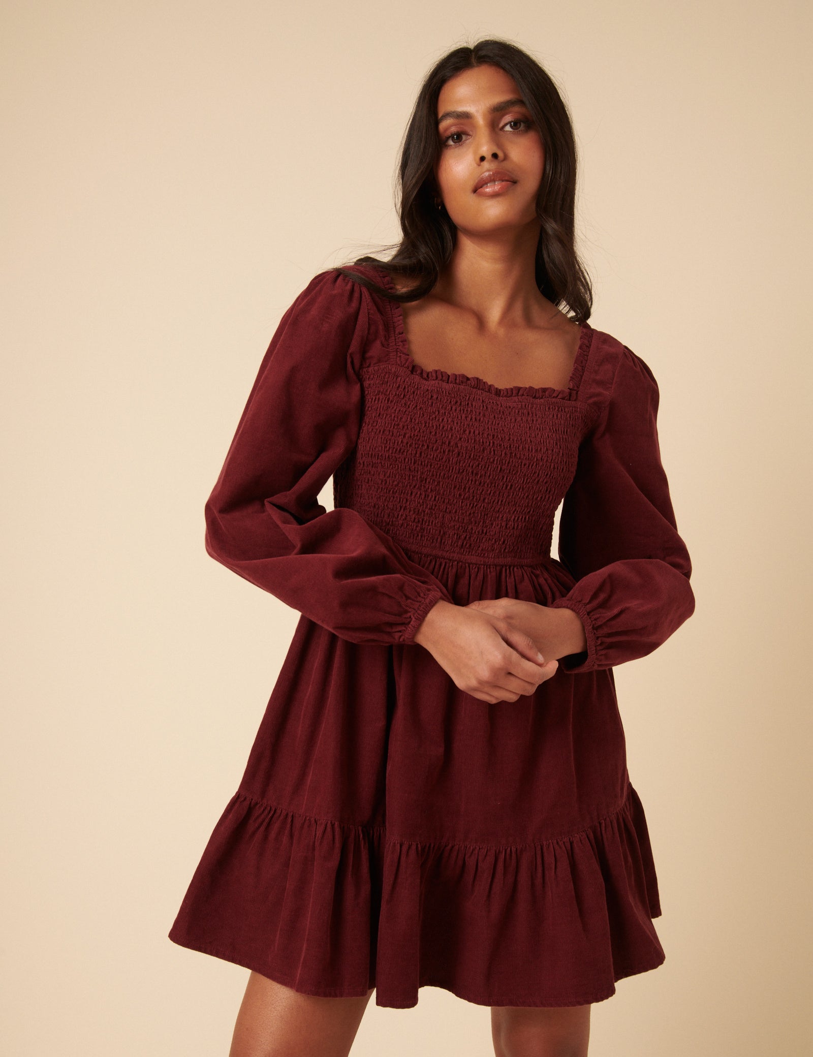 A woman wearing a dusty red mini dress with a square neckline, long balloon sleeves, shirred bodice and back, and a tiered, flowy hem.