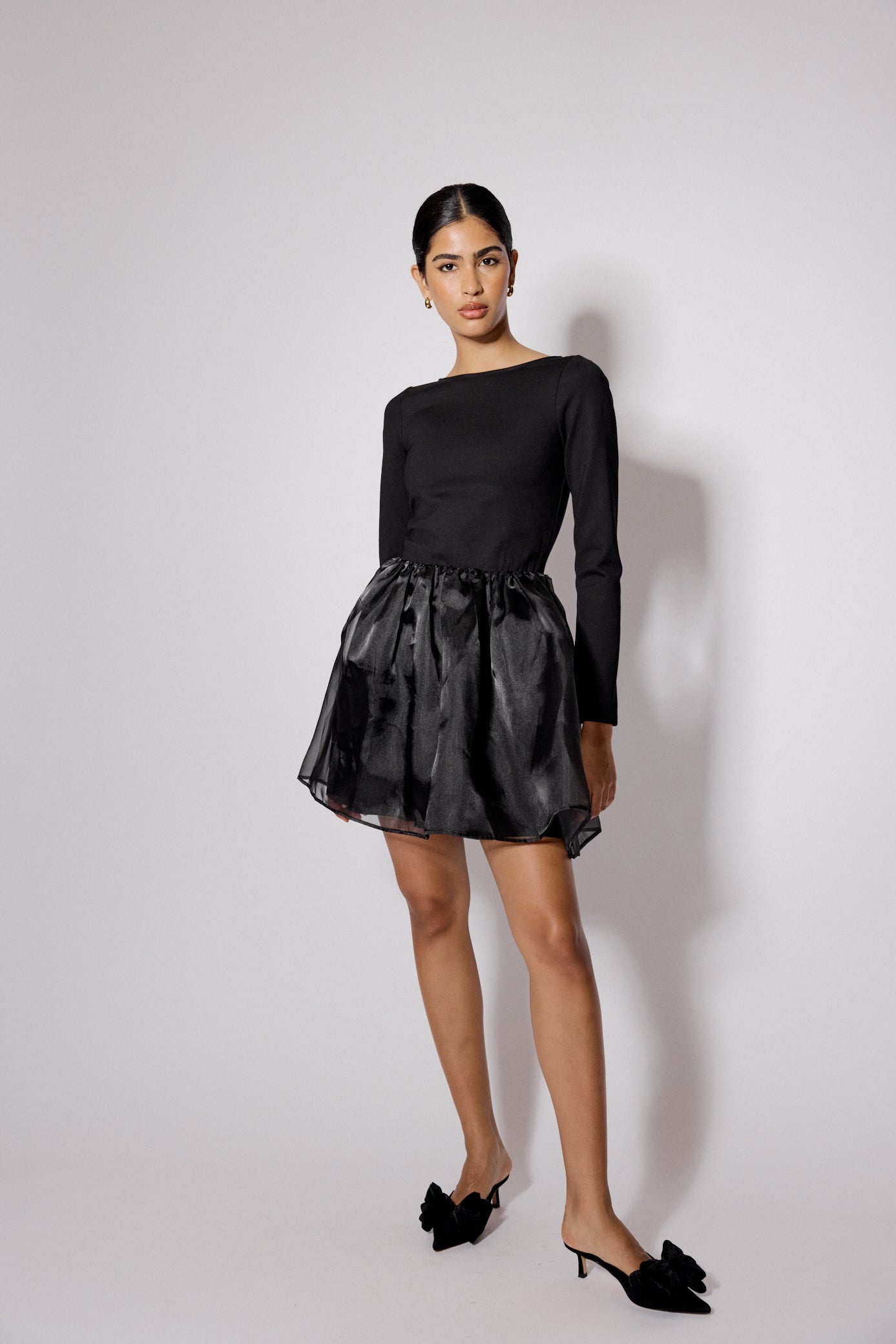 A woman wearing a black long sleeve organza mini dress with a boat neckline, fitted waist, and open back.