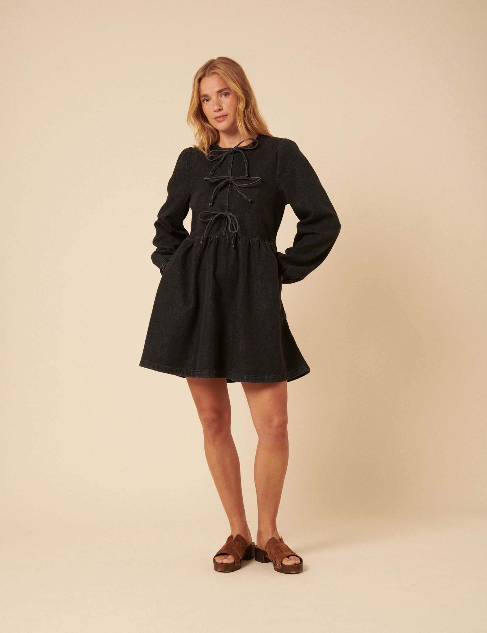 A woman wearing a black denim mini dress with long sleeves, a tie-front at the bodice, and side pockets.